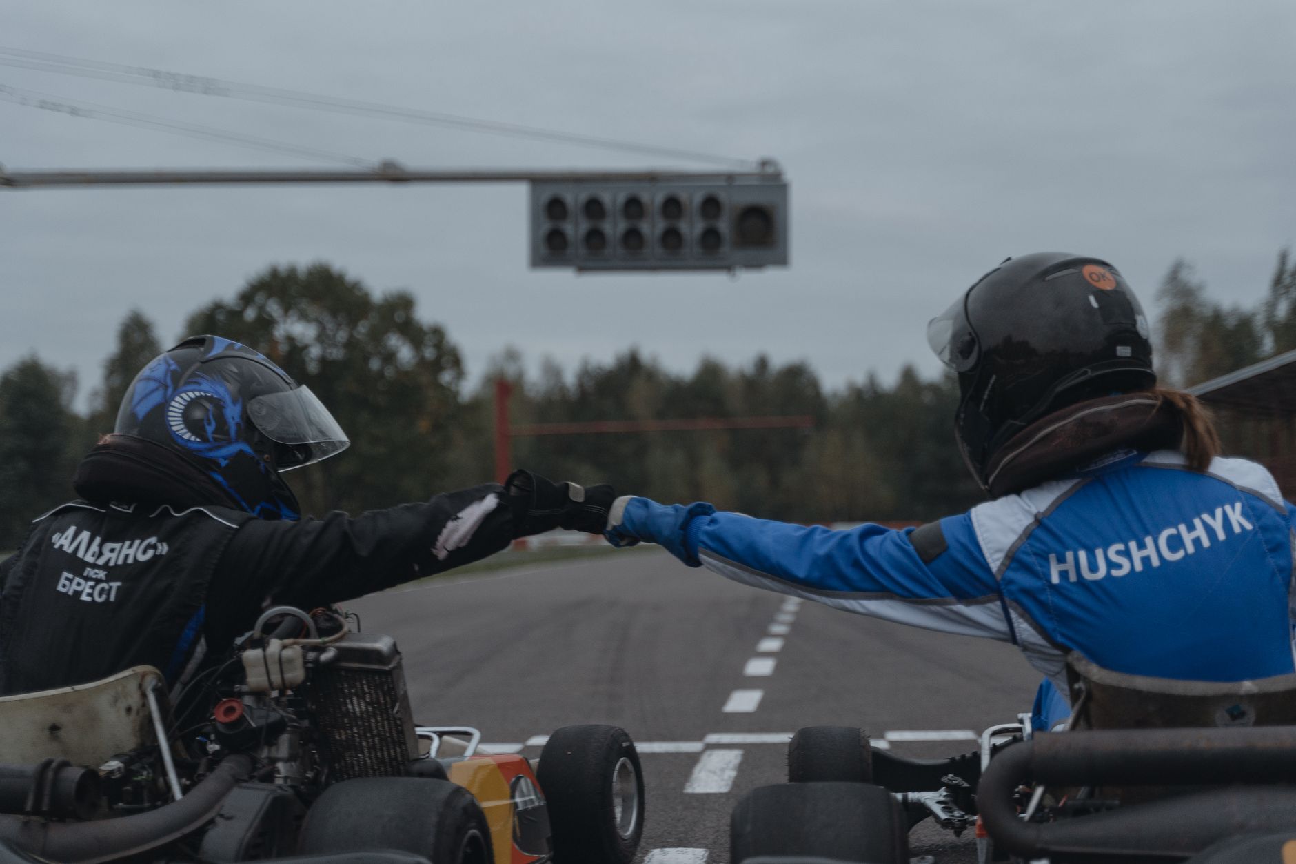 Dua pembalap kart bertukar fist bump di trek balap, menunjukkan semangat olahraga dan kerja tim.
