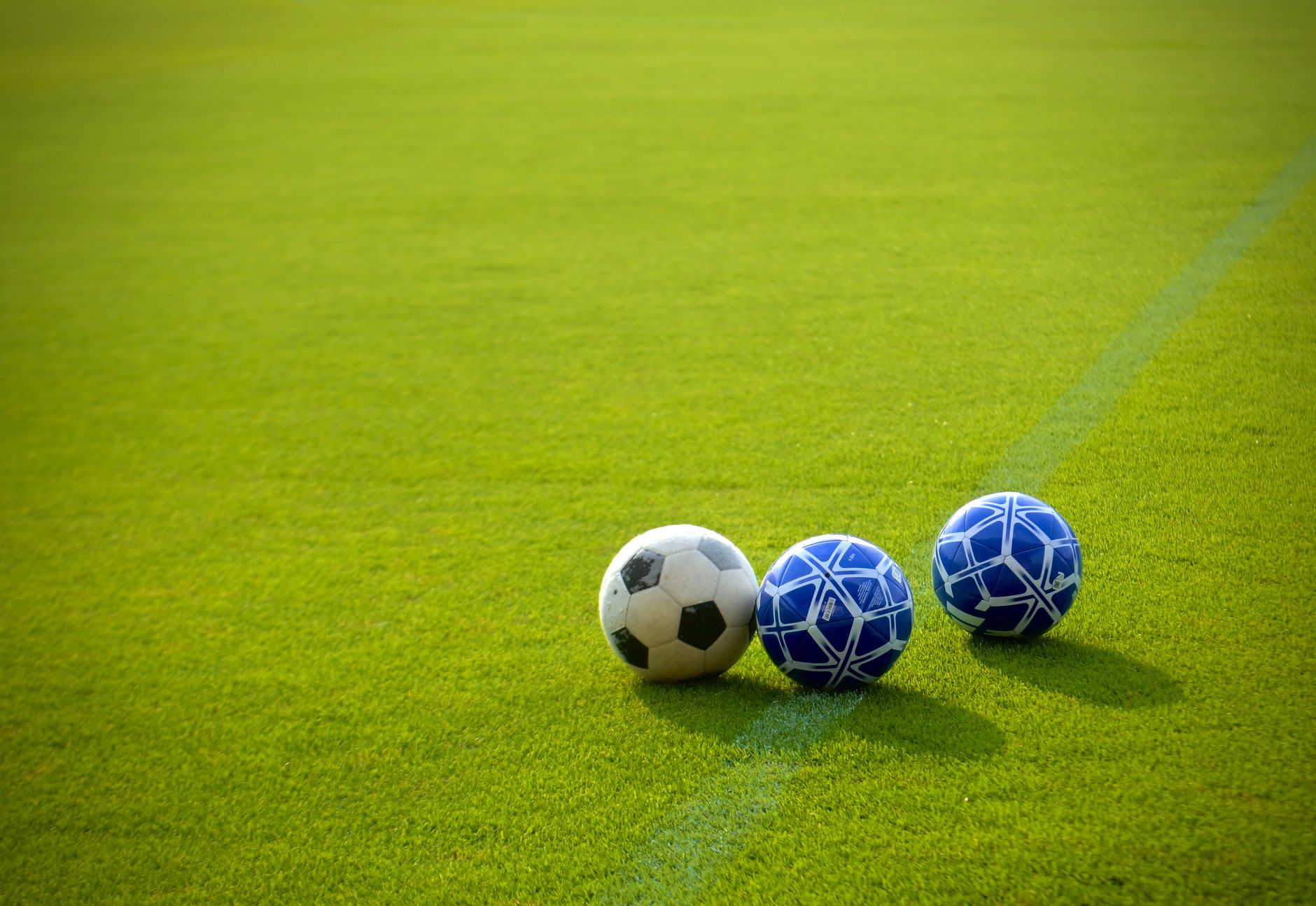 Tiga bola sepak ditempatkan di lapangan hijau yang cerah di bawah sinar matahari sore, sangat cocok untuk tema olahraga.