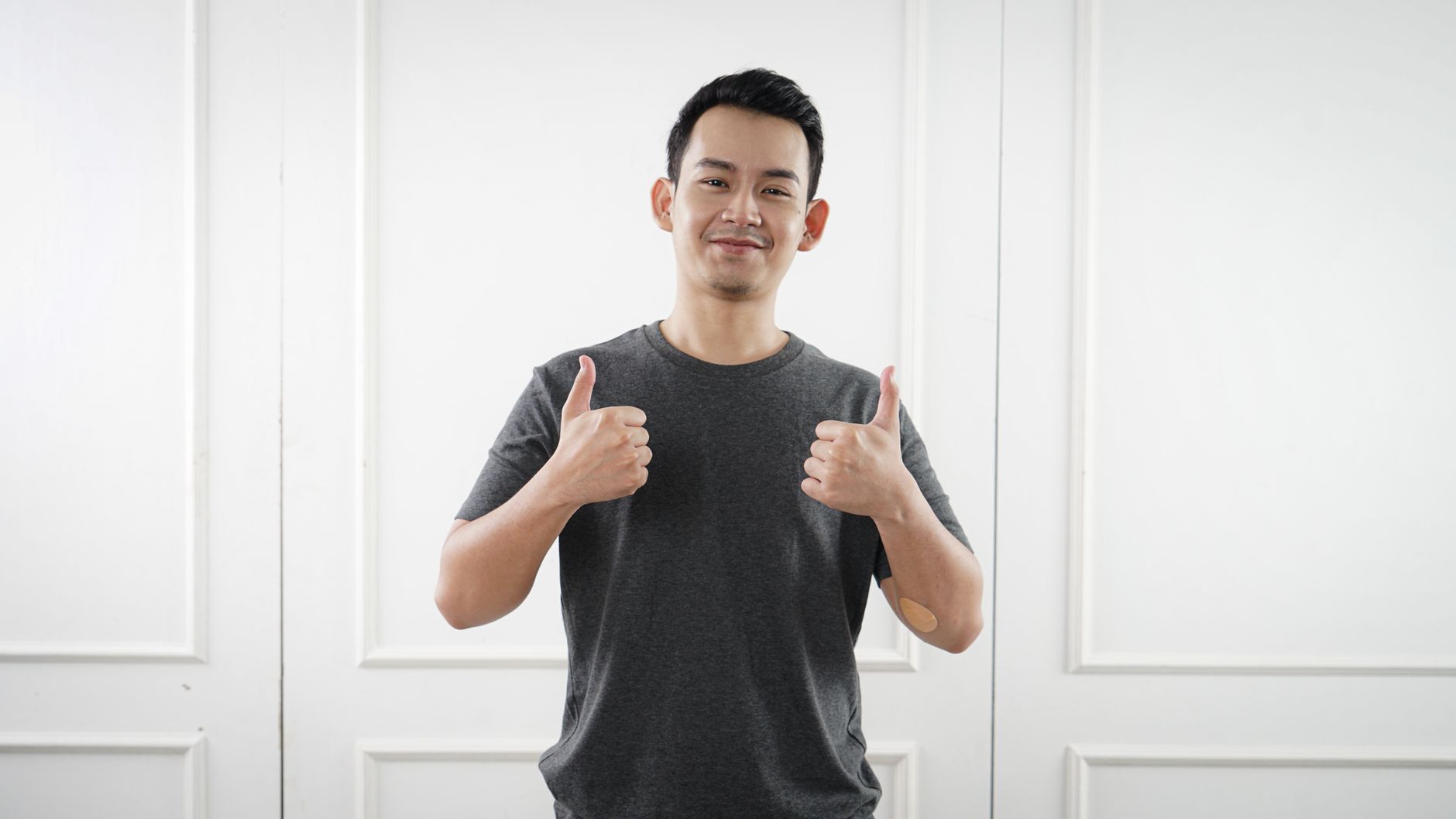 Pria Asia dalam kaos abu-abu menunjukkan jari telunjuk ke atas sambil tersenyum di dalam ruangan di Jakarta.