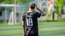 Pemain muda dengan jersey hitam di lapangan sepak bola luar ruangan pada siang hari, menghadap menjauh dari kamera.