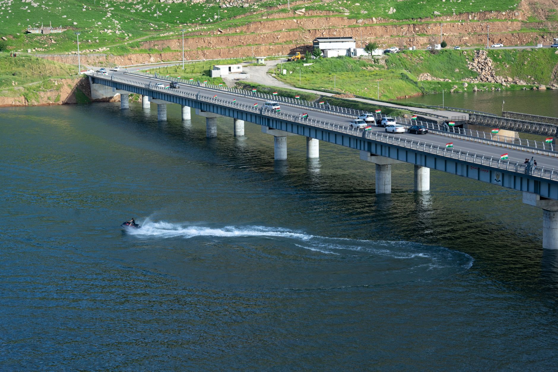 Sebuah jembatan membentang di atas danau biru di Kurdistan, dengan jet ski menciptakan jejak di atas air.