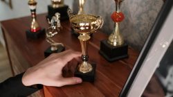 Sebuah foto close-up tangan yang menjangkau piala berkilau di rak kayu, melambangkan kesuksesan.