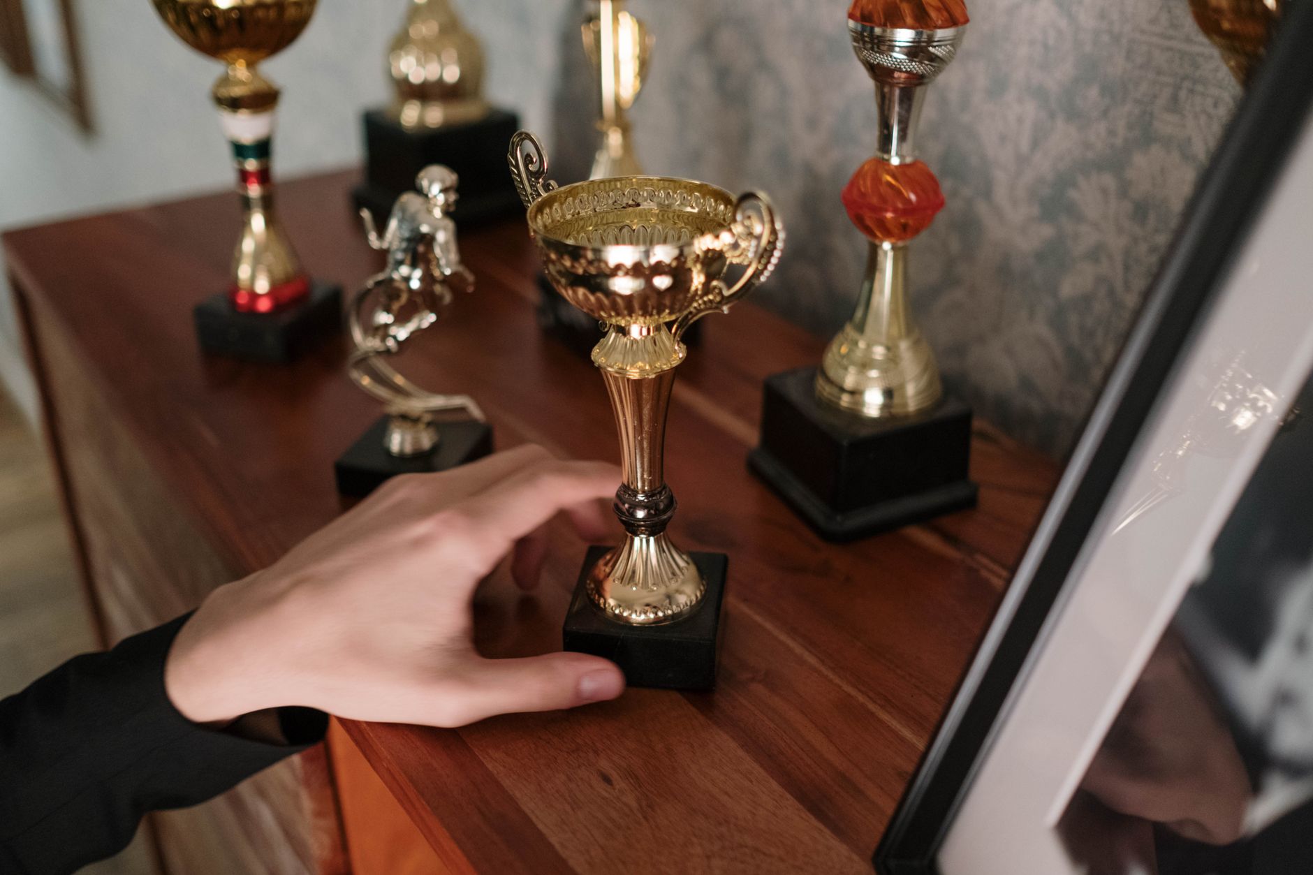 Sebuah foto close-up tangan yang menjangkau piala berkilau di rak kayu, melambangkan kesuksesan.