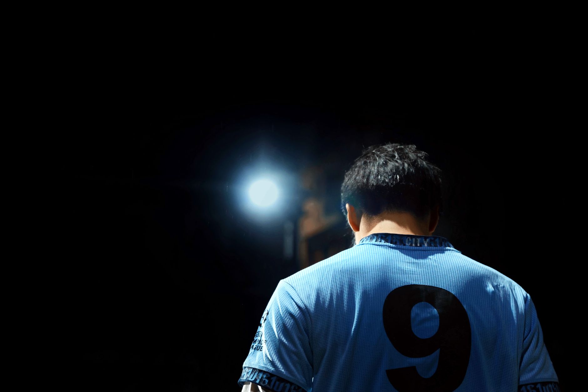 Seorang pemain sepak bola dengan jersey biru bernomor 9, diterangi oleh lampu sorot di malam hari.