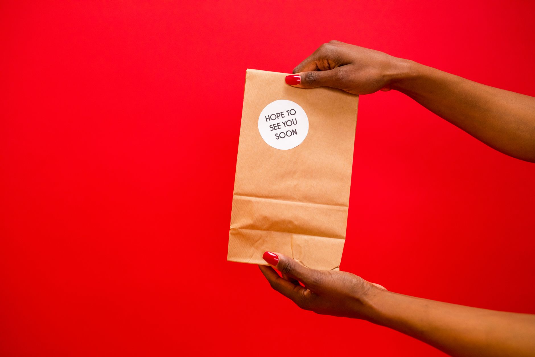 Dua tangan memegang kantong kertas dengan stiker 'Semoga segera bertemu', di latar belakang merah.