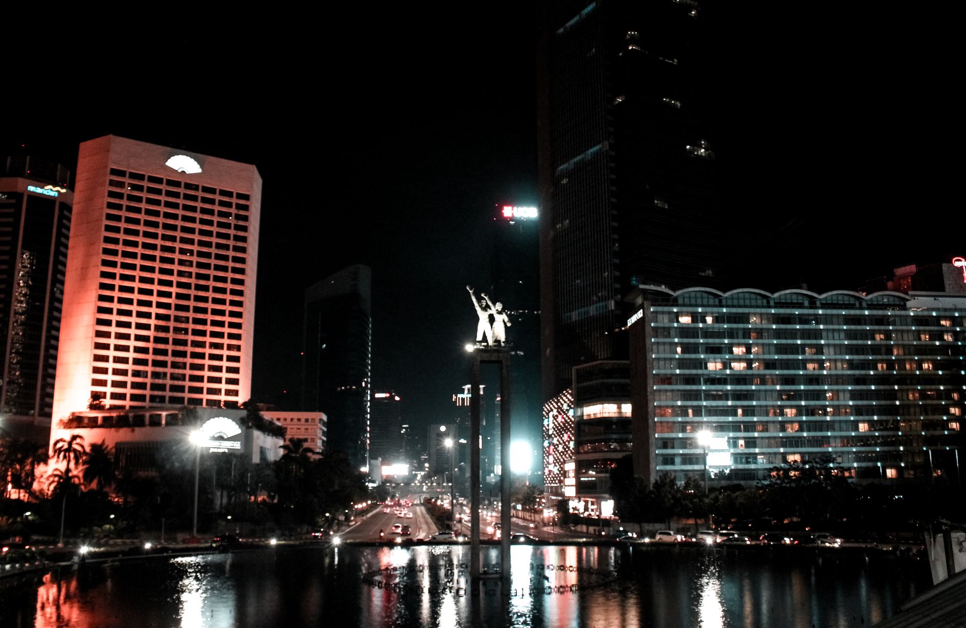 Monumen Selamat Datang yang diterangi dan gedung pencakar langit Jakarta di malam hari.