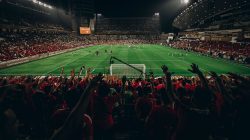 Pemandangan udara pertandingan sepak bola yang meriah di stadion yang penuh sesak pada malam hari, menangkap kegembiraan para penggemar olahraga.