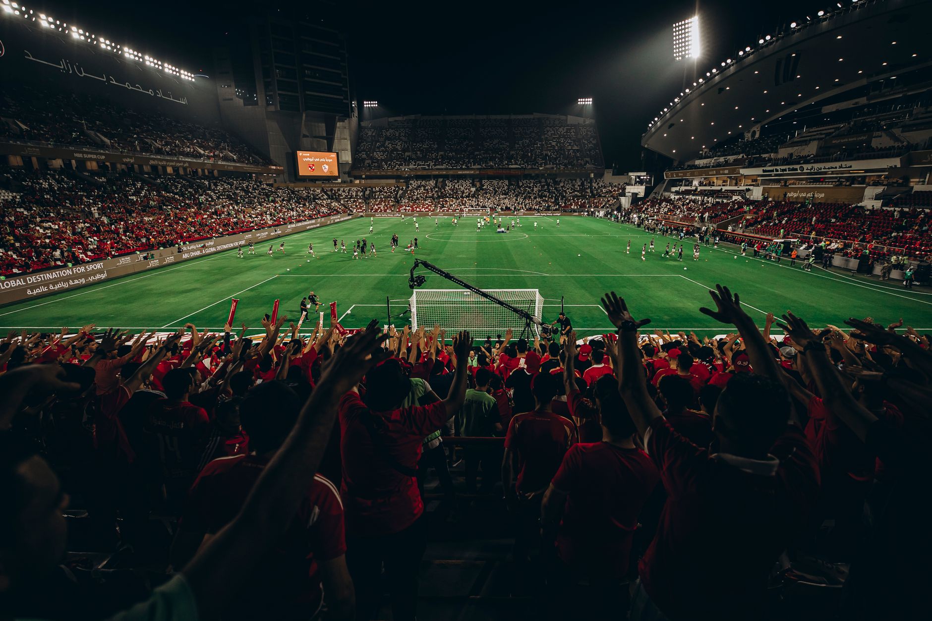 Pemandangan udara pertandingan sepak bola yang meriah di stadion yang penuh sesak pada malam hari, menangkap kegembiraan para penggemar olahraga.