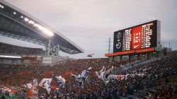 Sebuah adegan stadion sepak bola yang hidup dengan penggemar melambaikan bendera dan papan skor menampilkan rincian pertandingan.